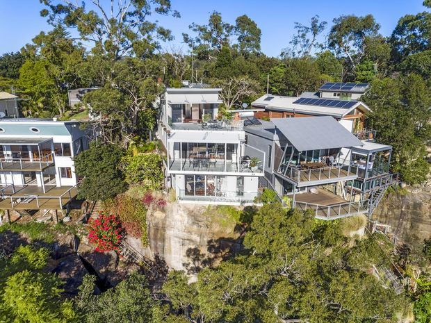 Aerial view of several modern residential homes built into a heavily vegetated, elevated and sloping site. The central multi-level home features extensive glazing, balconies and terraces, while the neighbouring homes have pitched and skillion-style roofs, timber decks and visible rooftop solar panels. Dense native-looking trees surround the properties.