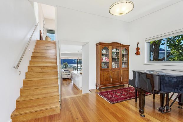 Bright open-plan interior featuring a timber staircase, polished timber flooring, a traditional wooden display cabinet, red patterned rug, and a black grand piano. A living area and large glazed opening are visible beyond the hallway, with a small garden view through the side window.