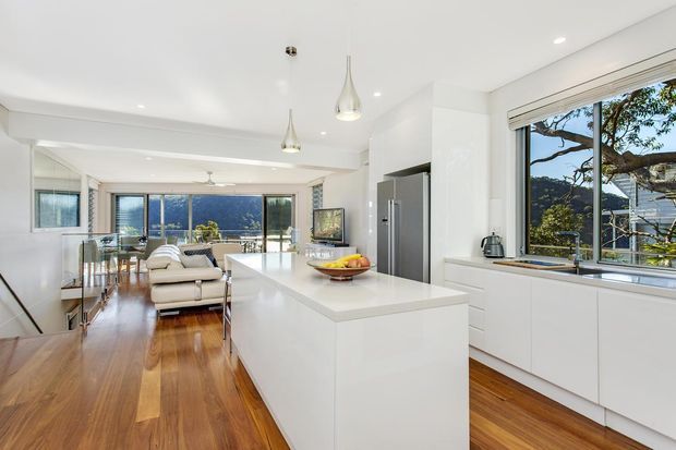 Bright open-plan kitchen with a large white island, integrated white cabinetry, stainless-steel refrigerator, pendant lights, and timber flooring. The kitchen opens directly to a dining and living area with wide glazing and views of leafy surroundings and water or hills beyond.