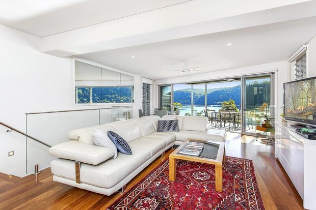Bright contemporary living room with a large white sectional sofa, patterned red area rug, timber coffee table, built-in wall-mounted television, and expansive sliding glass doors opening to a balcony with a scenic waterfront or bay view. White walls, recessed lighting, timber flooring, and a glass stair balustrade create an airy, open-plan feel.