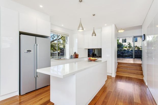 Bright contemporary white kitchen with a large white island, integrated white cabinetry, stainless-steel double-door refrigerator, pendant lights, and warm timber flooring. The kitchen opens toward a small raised timber-floored outdoor or balcony area through glazing and a doorway.
