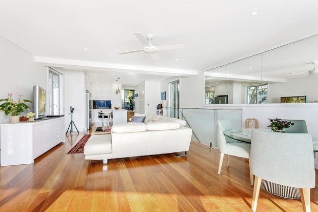 Bright open-plan living and dining area with white walls and built-in cabinetry, polished warm timber flooring, a white sectional sofa, glass dining table, upholstered dining chairs, large mirrored wall panels, recessed lighting, and a white ceiling fan. The room has abundant natural light and a clean contemporary coastal feel.