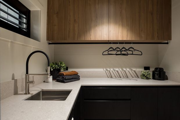 Compact, refined laundry room with dark lower cabinetry, a light stone benchtop, integrated stainless-steel sink and gooseneck tap, timber overhead cabinetry, hanging rail, and concealed upper ventilation window. Warm under-cabinet lighting and neatly folded towels create a polished, functional fit-out.