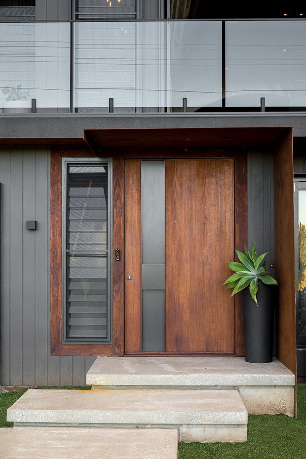 Close-up exterior view of a contemporary residential entry. A large warm timber pivot-style front door with narrow frosted-glass inserts and a matching timber-framed louvred sidelight is set within dark vertical cladding and a deep timber-lined entry surround. Concrete steps lead to the doorway, with a black planter and green foliage beside it. An upper-level glass balustrade is visible above.