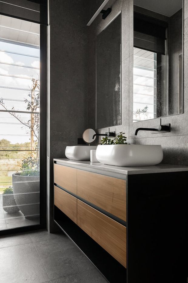 A contemporary bathroom vanity with two white vessel basins, matte black wall-mounted faucets, and a floating timber-fronted cabinet with dark framing. Large mirrors and floor-to-ceiling glazing bring in natural light, while dark grey textured wall and floor tiles create a refined, monochromatic atmosphere. Potted plants are visible near the glazing.