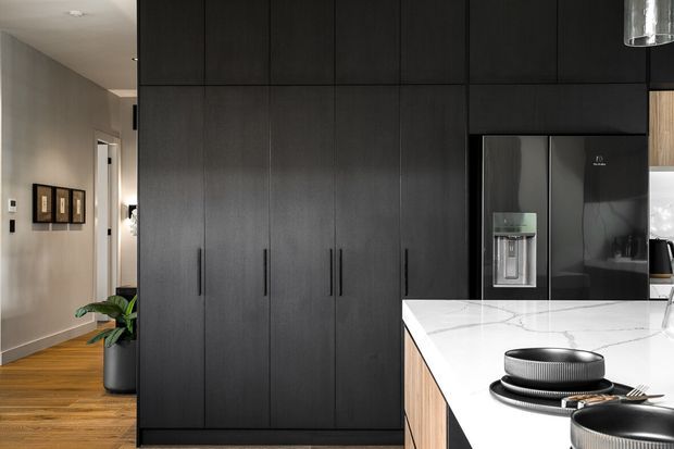 A contemporary kitchen viewed from an open-plan living or hallway area. The space features a large island with a white veined stone benchtop, dark integrated cabinetry extending floor to ceiling, a black refrigerator, timber island joinery, and warm timber flooring. A hallway with framed artwork and a potted plant is visible to the left.