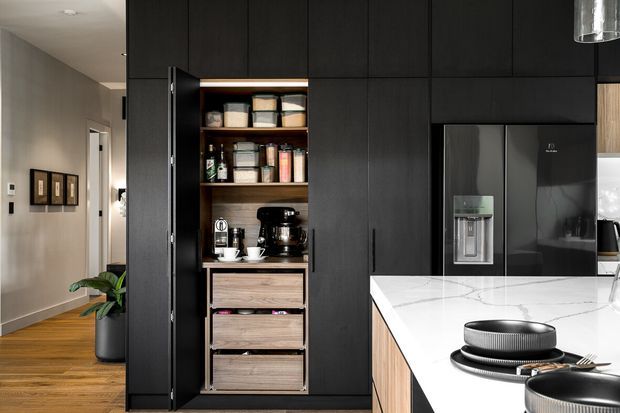 Contemporary kitchen featuring extensive matte black full-height cabinetry, an open timber-lined appliance and pantry niche, integrated black refrigerator, and a bright white stone island benchtop with a built-in cooktop. Warm timber flooring and neutral grey walls extend into the adjacent hallway.