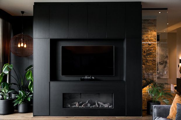 Moody contemporary living room centered on a full-height matte-black media wall with integrated television and linear fireplace. Dark built-in cabinetry contrasts with exposed brickwork, indoor plants, a woven pendant light, and warm timber flooring.