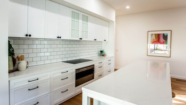 Bright, contemporary white kitchen with extensive shaker-style cabinetry, a white subway-tile splashback, black hardware, an integrated cooktop and oven, and a large glossy white island. Warm timber flooring and a framed artwork add contrast.