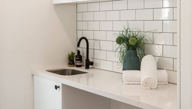 A bright, compact kitchenette or utility sink area with white shaker-style cabinetry, a white benchtop, black gooseneck tapware, stainless-steel undermount sink, and a white subway-tile splashback with dark grout. Decorative plants, soap dispensers, and rolled towels sit on the counter.