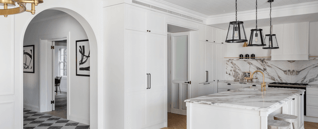 Bright, refined white kitchen with extensive built-in cabinetry, a large marble-look island and matching splashback, brass tapware, black-framed pendant lights, and a black-and-white checkerboard floor visible through the arched opening. The space connects visually to a hallway and adjoining rooms.