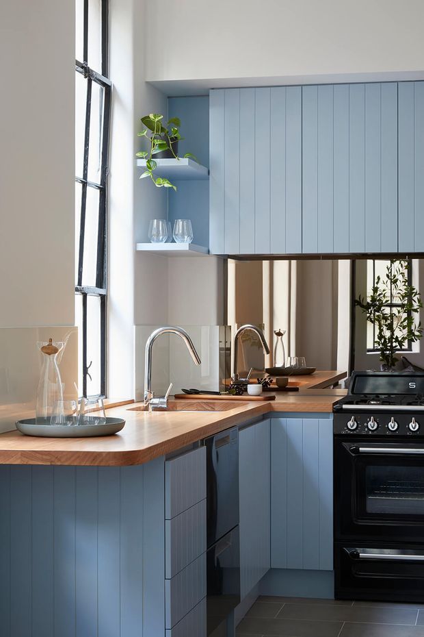 A bright, compact kitchen with pale blue vertical-panel cabinetry, warm timber benchtops, a black-framed industrial-style window, stainless steel gooseneck taps, mirrored splashback, open shelving, and a black freestanding double oven. A small plant and glassware add restrained decorative detail.