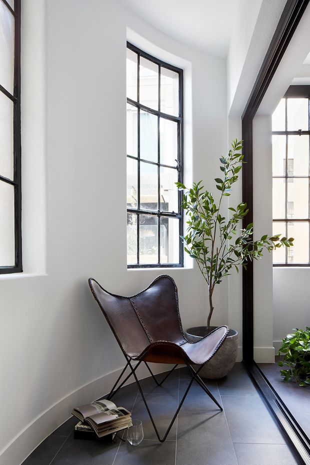 Bright, narrow interior nook with white walls, tall black-framed industrial-style windows, dark tiled flooring, and a dark leather sling chair beside a potted indoor tree. A large glazed opening connects visually to an adjacent bright space, while natural light creates an airy, refined atmosphere.