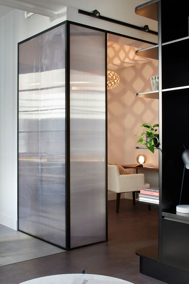 A contemporary interior featuring a black-framed translucent sliding partition separating a bright tiled area from a warm, softly lit lounge or living space. The lounge includes a cream upholstered armchair, dark timber flooring, a small side table, decorative pendant lighting, indoor plants, and integrated black shelving with curved timber-edged shelves. The translucent partition and patterned light shadows create a layered, refined atmosphere.