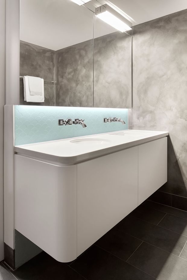 A contemporary bathroom vanity viewed straight on, featuring a floating white double-basin unit with integrated sinks and wall-mounted chrome faucets. Behind it is a pale aqua scallop-pattern tiled splashback, while the surrounding walls are finished in mottled grey plaster or concrete-look coating. A large mirrored cabinet spans the vanity wall, and dark rectangular floor tiles contrast with the bright white cabinetry. A towel rail and folded towel are visible at left, with linear ceiling lighting above.