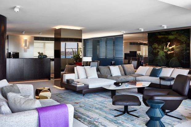 A sophisticated open-plan living room with a large modular sectional sofa, lounge chairs, ottoman, and low coffee table arranged over a patterned rug. Dark timber or stained built-in cabinetry, concealed storage, mirrored columns, white ceilings, and dark blue plantation shutters create a refined contemporary interior. A large dramatic artwork forms a feature wall, while layered lighting and expansive glazing bring in natural light.
