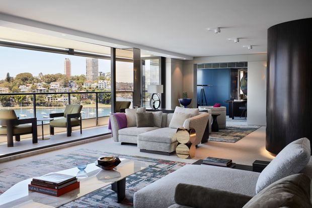 A sophisticated contemporary living room with expansive floor-to-ceiling glazing overlooking a riverfront or lakeside urban landscape. The room features a large pale sectional sofa, sculptural lounge chairs, layered patterned rugs, a low white coffee table, dark timber or black accents, and a dramatic curved dark feature wall or column. Blue cabinetry and artwork add colour in the background, while abundant natural light creates an airy, refined atmosphere.