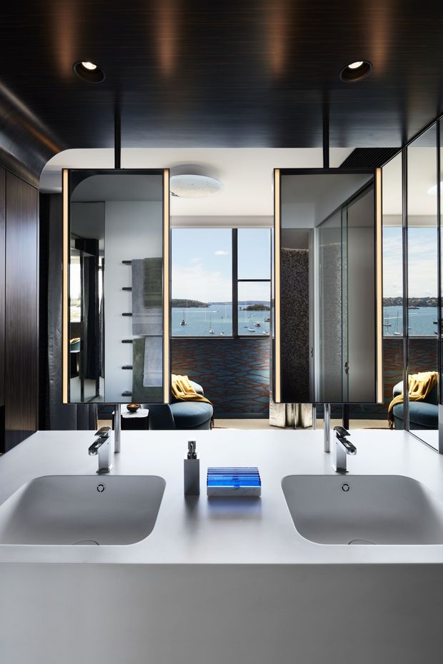 A contemporary double-vanity bathroom viewed toward a large window overlooking a blue waterfront. The room features a white integrated vanity with two sculpted basins and chrome taps, mirrored cabinetry, dark vertical wall panelling, a dark ceiling with recessed spotlights, glass shower enclosures, and a patterned dark feature wall. Reflections reveal additional bathroom fittings and a dark upholstered chair.