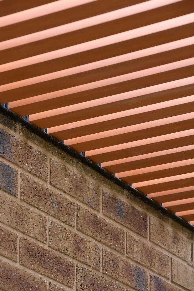 Close-up architectural detail of a textured brown brick wall meeting an overhead pergola or slatted roof. Parallel warm terracotta-brown beams create a strong linear pattern, with narrow gaps allowing light through. The image focuses on exterior construction materials and structural detailing, with no furnishings or room interior visible.