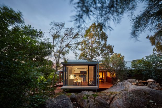 A compact modern glass-fronted pavilion or house extension nestled among dense native vegetation and large exposed rock formations. The dark-framed glazing reveals a warmly lit interior, while a timber-toned adjoining volume and deck extend to the right. The image is taken from outdoors at dusk, emphasizing the building’s integration with its rugged natural setting.