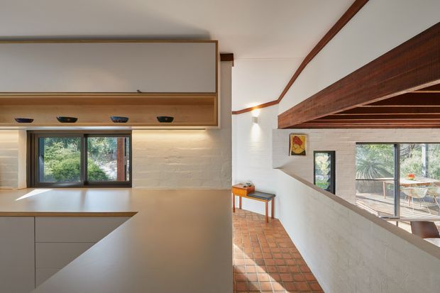 Interior view of a warm, contemporary split-level living space with an integrated kitchen or joinery wall in the foreground. White textured masonry walls, extensive dark timber beams and trim, timber cabinetry, and terracotta tiled flooring define the space. A low internal opening overlooks a dining or living area with large windows, while greenery is visible outside.
