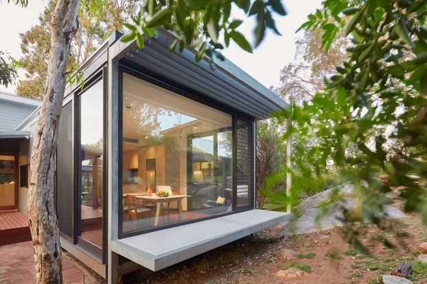 Exterior view of a compact contemporary glazed extension set among established trees and rocky landscaped ground. The dark-framed floor-to-ceiling glazing reveals a warm dining area inside, while a projecting flat roof with exposed ribbed soffit and a pale concrete-like ledge define the modern addition. A timber deck and adjoining older structure are visible to the left.