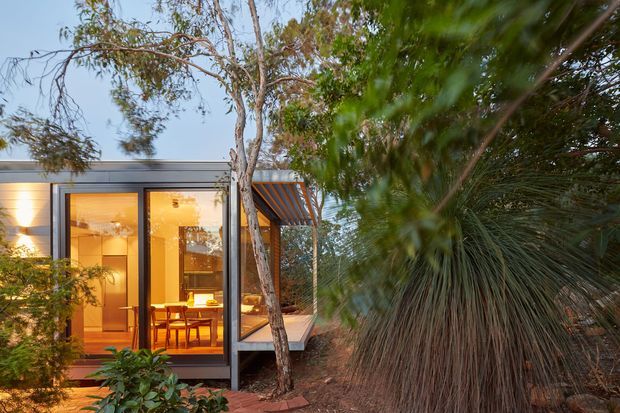 Exterior view of a contemporary single-storey residence set within dense landscaped vegetation. The dark, low-profile facade has extensive floor-to-ceiling glazing revealing a warmly lit dining area, with a projecting timber-lined roof or awning and a small deck or paved terrace at the entrance. Mature trees and ornamental grasses frame the building.