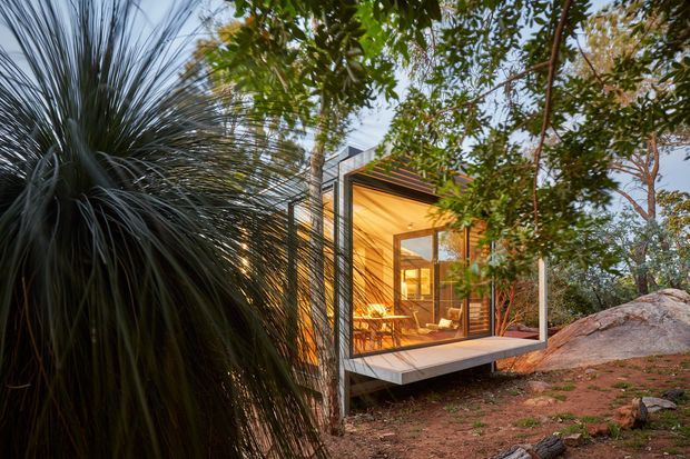 A compact modern glass-fronted cabin or pavilion nestled among dense native vegetation and exposed rock. The structure has a projecting, elevated box form with dark metal framing, extensive glazing, warm timber-lined interior surfaces, and a flat or very low-pitched roof. A small dining or living area is visible inside, while the surrounding bushland forms a natural, secluded setting.