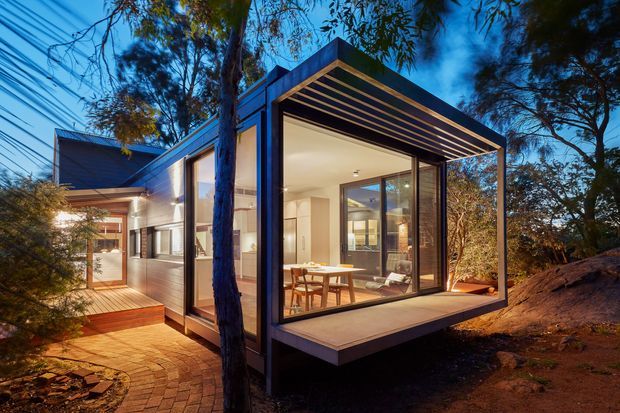 A contemporary compact home extension or pavilion set among mature trees, photographed at dusk. The dark framed rectangular structure features extensive floor-to-ceiling glazing, a projecting flat roof with exposed linear soffit battens, and a raised deck-like platform. Through the glazing, a bright dining and living area is visible. A brick-paved walkway connects to the adjoining building, with warm interior and exterior lighting creating an inviting atmosphere.