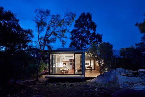A compact modern single-storey home or pavilion photographed at dusk, set among mature trees and exposed rocky terrain. Floor-to-ceiling glazing reveals a warmly lit dining and living area, while a covered or adjoining terrace extends to the right. The building has a dark, minimalist rectilinear form with a low flat roof and strong indoor-outdoor connection.