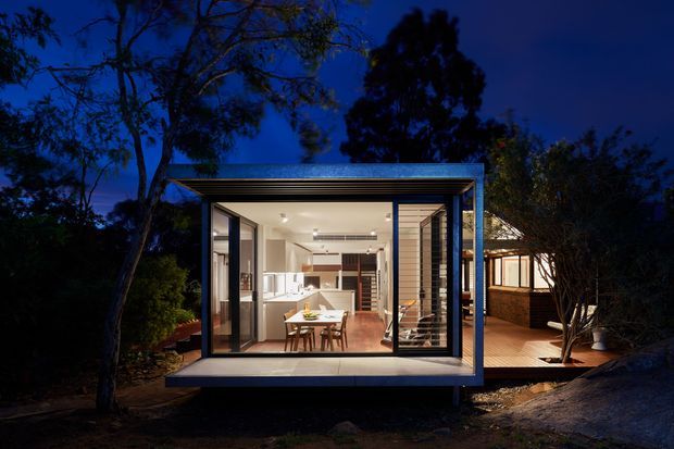 Night-time exterior view of a compact contemporary glass-fronted pavilion or extension set within a landscaped residential site. The structure has a flat roof with a projecting box-like form, dark metal-framed glazing, pale wall surfaces, and a timber deck extending to the side. A brightly lit open-plan kitchen and dining area is visible through the glazing, with adjacent residential structures and mature trees in the background.