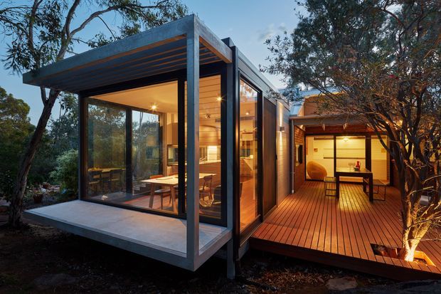 A modern glass-walled pavilion-style home set among mature trees, with a flat-roofed concrete and dark-framed glazed volume opening onto a warm timber deck. The illuminated interior appears to contain a dining area and kitchen, while a connected covered outdoor space includes a table and seating. The design has strong indoor-outdoor connections, exposed structural framing, and a warm evening atmosphere.