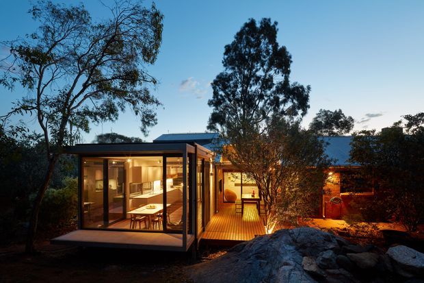 Twilight exterior view of a contemporary single-storey house set among mature trees and rocky landscaping. The home features extensive floor-to-ceiling glazing, dark slender frames, warm timber decking, and warmly illuminated interior and outdoor living spaces. A low-pitched roof and connected pavilion-like volumes create a horizontal, integrated form within the natural setting.