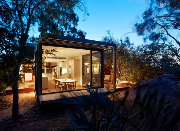 A small contemporary glass-walled pavilion or extension nestled among mature trees and large natural rocks, photographed at dusk. Warm interior lighting reveals an open-plan dining and living area with timber flooring, a dining table, staircase, and built-in joinery. The structure has dark slender framing, broad glazing, and a flat roof, with a timber deck and pathway connecting it to the surrounding landscape.