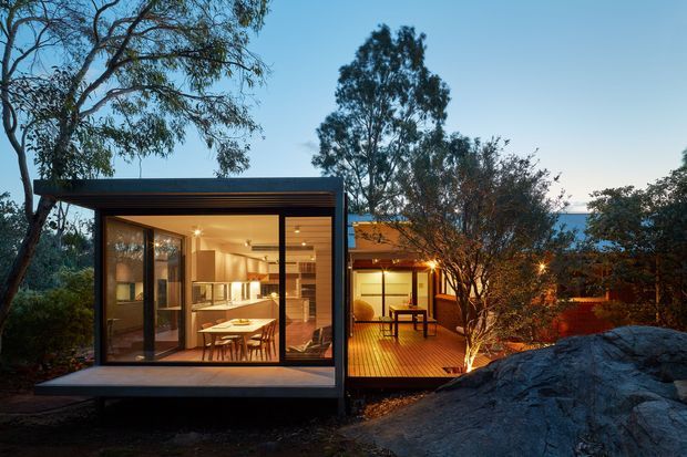Twilight exterior view of a compact contemporary dwelling set among mature trees and natural rock outcrops. The glazed pavilion-like volume has a dark framed facade, warm timber-toned interior lighting, and an open deck connecting to a smaller timber-clad section. The image shows strong indoor-outdoor connection and extensive glazing.