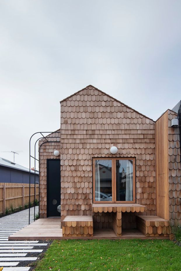 Exterior view of a compact contemporary dwelling clad almost entirely in overlapping rounded timber shingles, with a steep gabled facade, timber-framed window, dark entry door, integrated built-in bench seating, and a small timber deck. A striped paved path, timber boundary fence, lawn, and neighbouring structures are visible around the building.