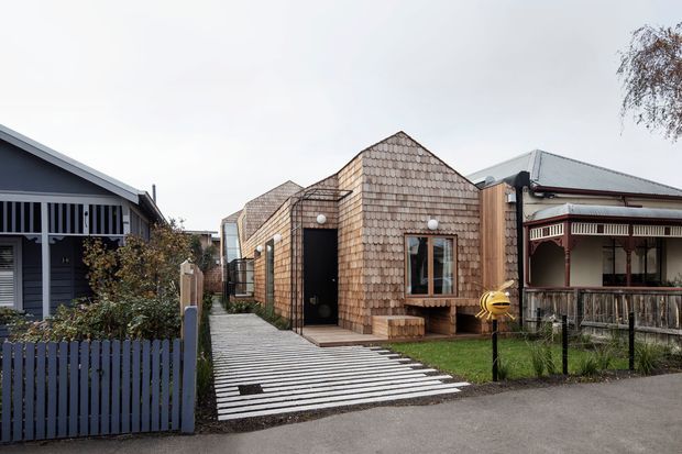 Street-level view of a compact contemporary residential building clad in overlapping timber shingles, positioned between neighbouring homes. The narrow frontage includes a gabled roof, dark entry door, timber-framed window, and a striped concrete or timber-look driveway/walkway. Traditional verandah housing is visible to the right and a darker weatherboard home with a picket fence to the left.