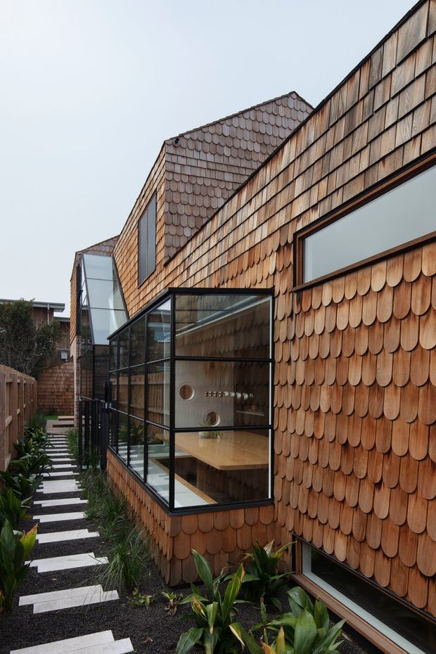 Exterior view of a contemporary multi-level residence clad in overlapping timber shingles, with black-framed glazing and a projecting glass-enclosed dining or living space. A narrow landscaped side path with rectangular stepping stones runs alongside the building.