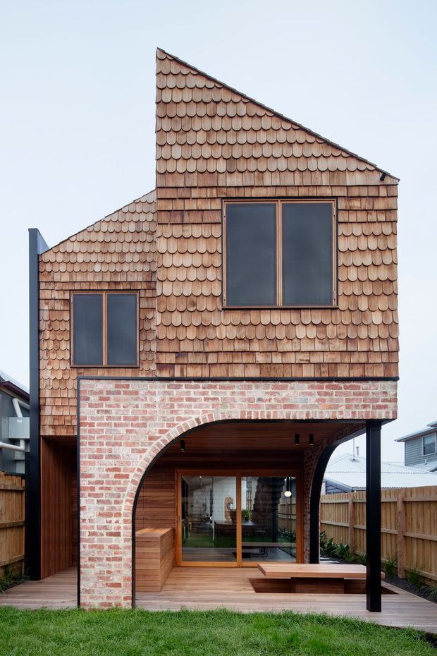 Front exterior view of a narrow multi-storey townhouse or residential extension. The upper facade is clad in overlapping timber shingles with large dark-framed windows, while the lower level features a pale red brick archway supported by dark steel posts. A timber-lined outdoor passage or covered patio with glazed doors sits beneath the arch, opening toward a fenced garden. The design combines strongly textured natural materials with a contemporary, compact urban form.