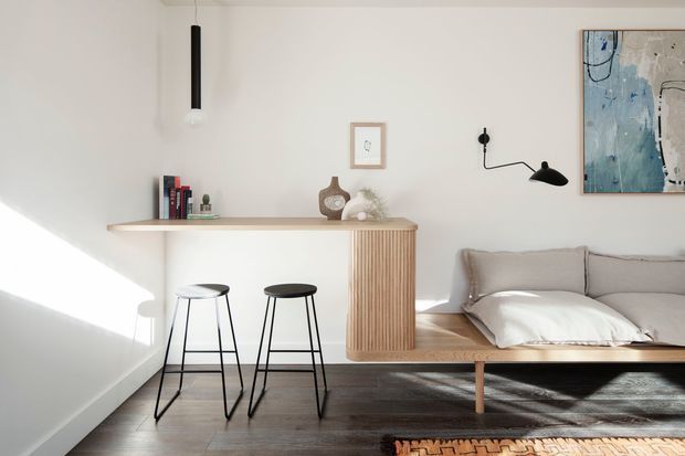 A bright, compact contemporary studio-like living and sleeping area with a built-in timber slatted joinery unit functioning as a shelf, counter, and bedside cabinet. Two black bar stools sit beneath the floating counter, while a low timber-framed bed with neutral bedding occupies the right side. White walls, dark timber flooring, minimalist pendant and wall lighting, and abstract artwork create a clean, warm minimalist interior.