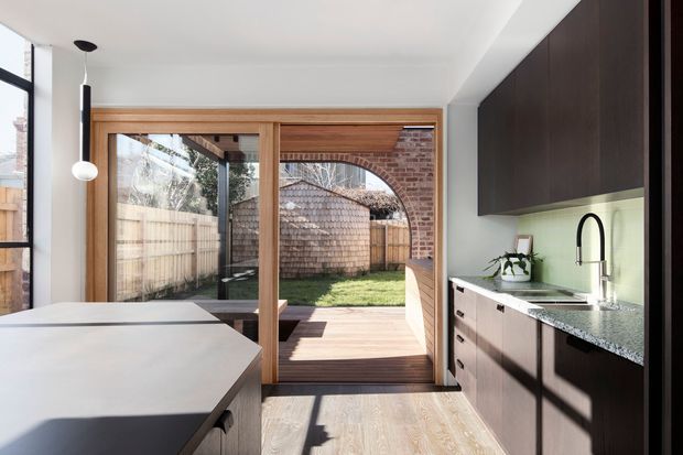 A bright contemporary kitchen opening onto a timber deck and enclosed backyard through large timber-framed sliding glass doors. The room features dark wood cabinetry, a pale stone benchtop, integrated under-cabinet lighting, a black gooseneck tap, light timber flooring, white walls, and a kitchen island with pendant lighting. Exposed brickwork and timber fencing are visible outside.