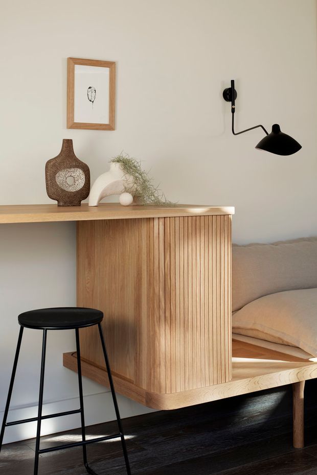 A warm, minimalist interior vignette featuring a light timber fluted built-in cabinet and counter integrated beside a cushioned timber-framed bench or daybed. A black wall-mounted articulated lamp and black metal stool contrast with the pale plaster wall and dark timber floor. Neutral ceramics, a framed print, and soft beige upholstery add understated styling.