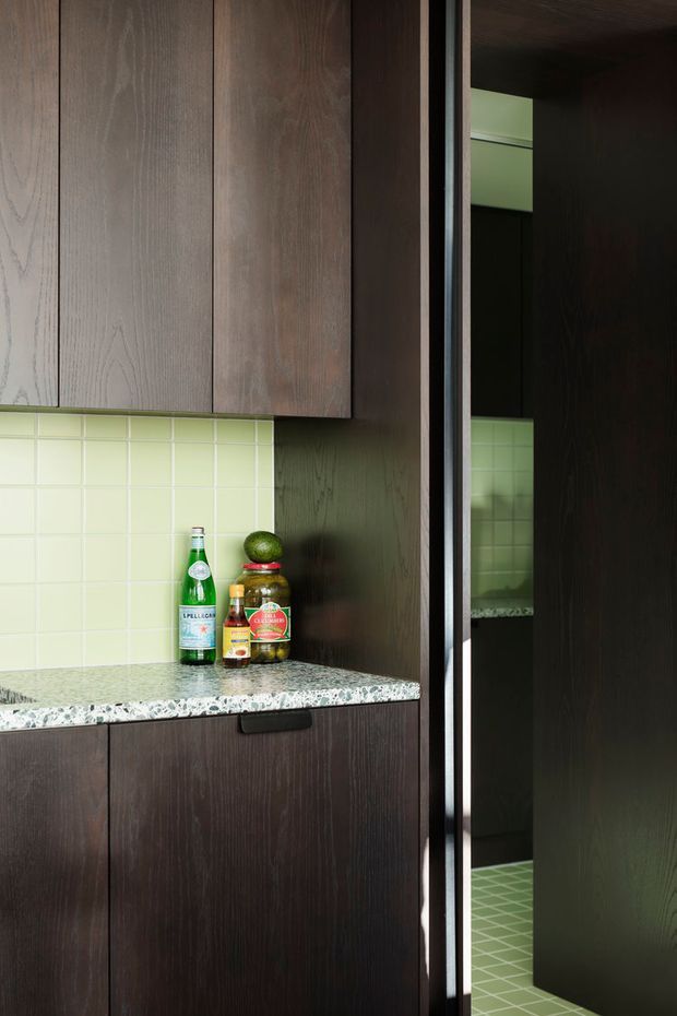 Close-up view of a compact, dark timber-toned kitchen or kitchenette with full-height wood-grain cabinetry, a pale green square-tile splashback, and a terrazzo-style stone benchtop. A tall dark cabinet or doorway at right includes a reflective surface, showing more green tiled surfaces beyond. Bottles and jars sit on the benchtop.