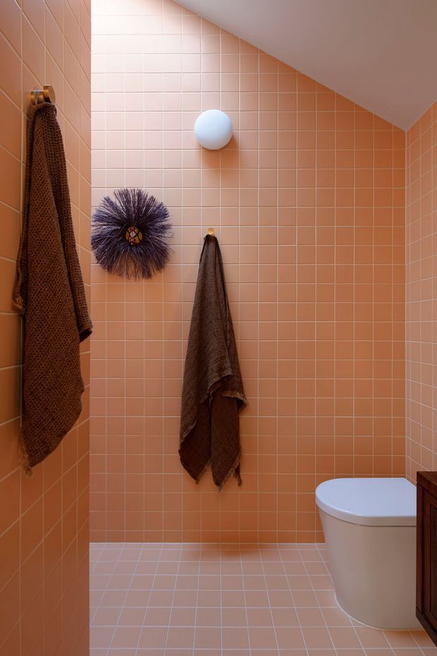 A compact bathroom with peach-pink square tiled walls and floor, a sloped ceiling, a round white wall light, hanging brown towels, decorative wall art, and a partially visible white toilet with dark timber-toned cabinetry. The space has a warm, playful, monochromatic tiled finish.