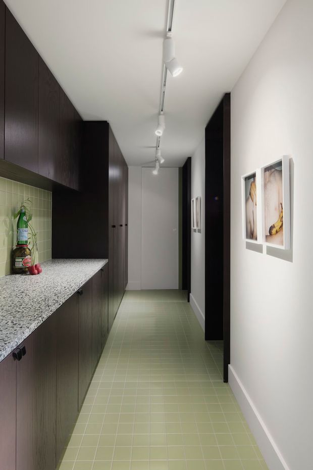 A narrow, modern galley kitchen and hallway viewed lengthwise. The left side has tall dark timber cabinetry, a speckled stone benchtop, and a pale green tiled splashback with a small sink area. The floor is finished in matching pale green square tiles. White walls and ceiling are illuminated by a linear track-lighting system, while framed artwork and dark built-in storage line the right side.