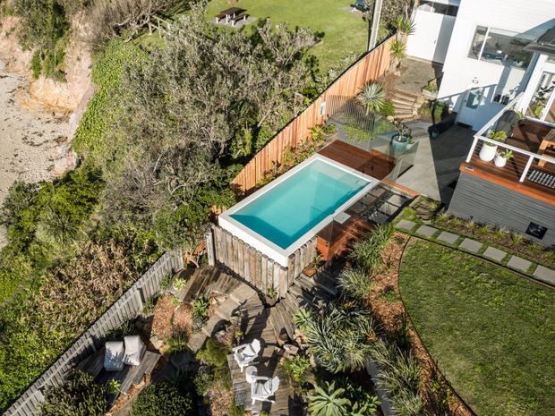 Elevated view of a contemporary residential outdoor area featuring a compact rectangular swimming pool, timber decking, paved paths, planted garden beds, lawn, timber fencing, and a white-clad house with outdoor stairs and terrace.