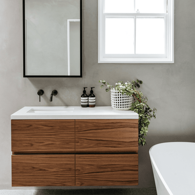 A refined modern bathroom featuring a floating walnut vanity with four wide drawers, a white integrated benchtop basin, matte black wall-mounted tapware, and black-framed mirror. The space has soft grey walls, a white-framed window, dark flooring, a freestanding white bathtub, and a trailing plant adding an organic accent.