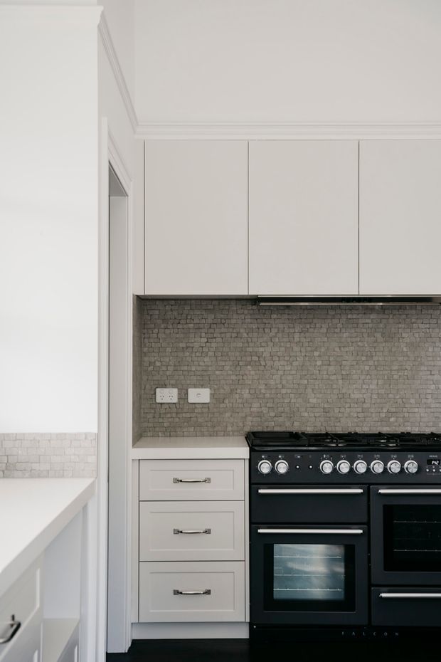 A narrow, refined kitchen composition featuring pale built-in cabinetry, a large black freestanding range cooker, and a grey textured brick-pattern splashback. Classic moulded cornices and trim contrast with the contemporary dark appliances, while a partial adjoining white counter and tiled wall are visible at left.