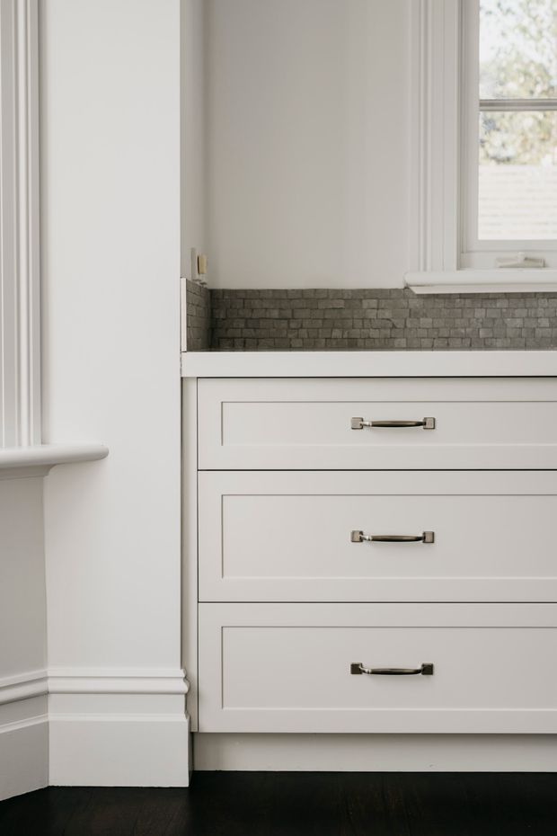 Close-up interior view of a built-in white painted drawer cabinet beneath a dark grey textured brick-tile backsplash. The cabinetry has recessed panel fronts with polished metal pulls, framed by white trim and moulding. A white-framed window and deep dark timber floor are visible, creating a refined, traditional-style fit-out.