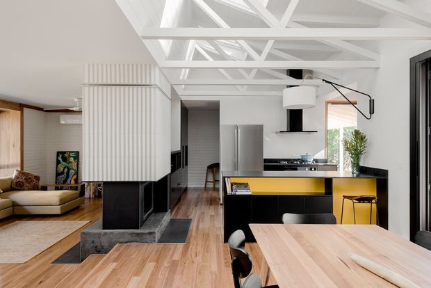 Bright open-plan residential interior combining a kitchen, dining area and living room. The space features white exposed roof trusses and a high ceiling, black lower cabinetry, a stainless-steel refrigerator, a dark island with a vivid yellow accent panel, light timber flooring, and a timber dining table. A compact living area is visible to the left, separated by a sculptural ribbed white partition and black fireplace or joinery element. Large glazing and skylight-like roof openings contribute to the airy atmosphere.